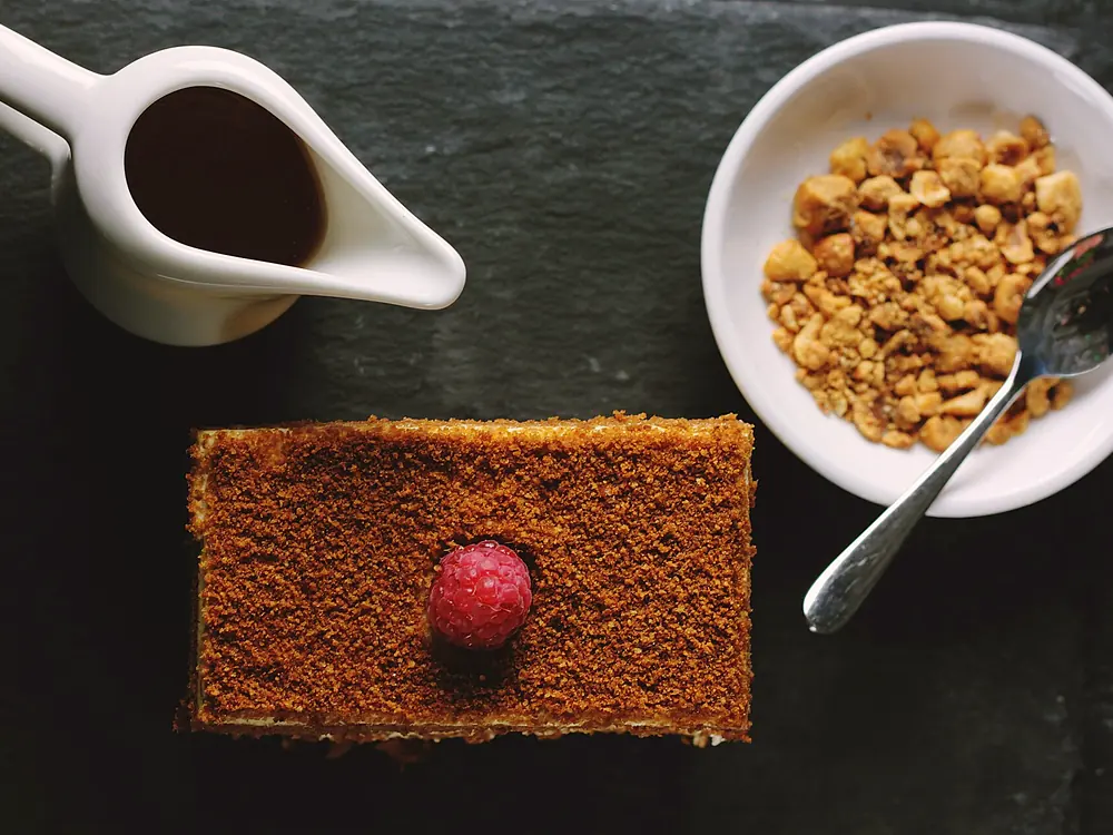 Fetta di dolce al cucchiaio su ardesia scura, con salsiera di cioccolato e granella di nocciole.