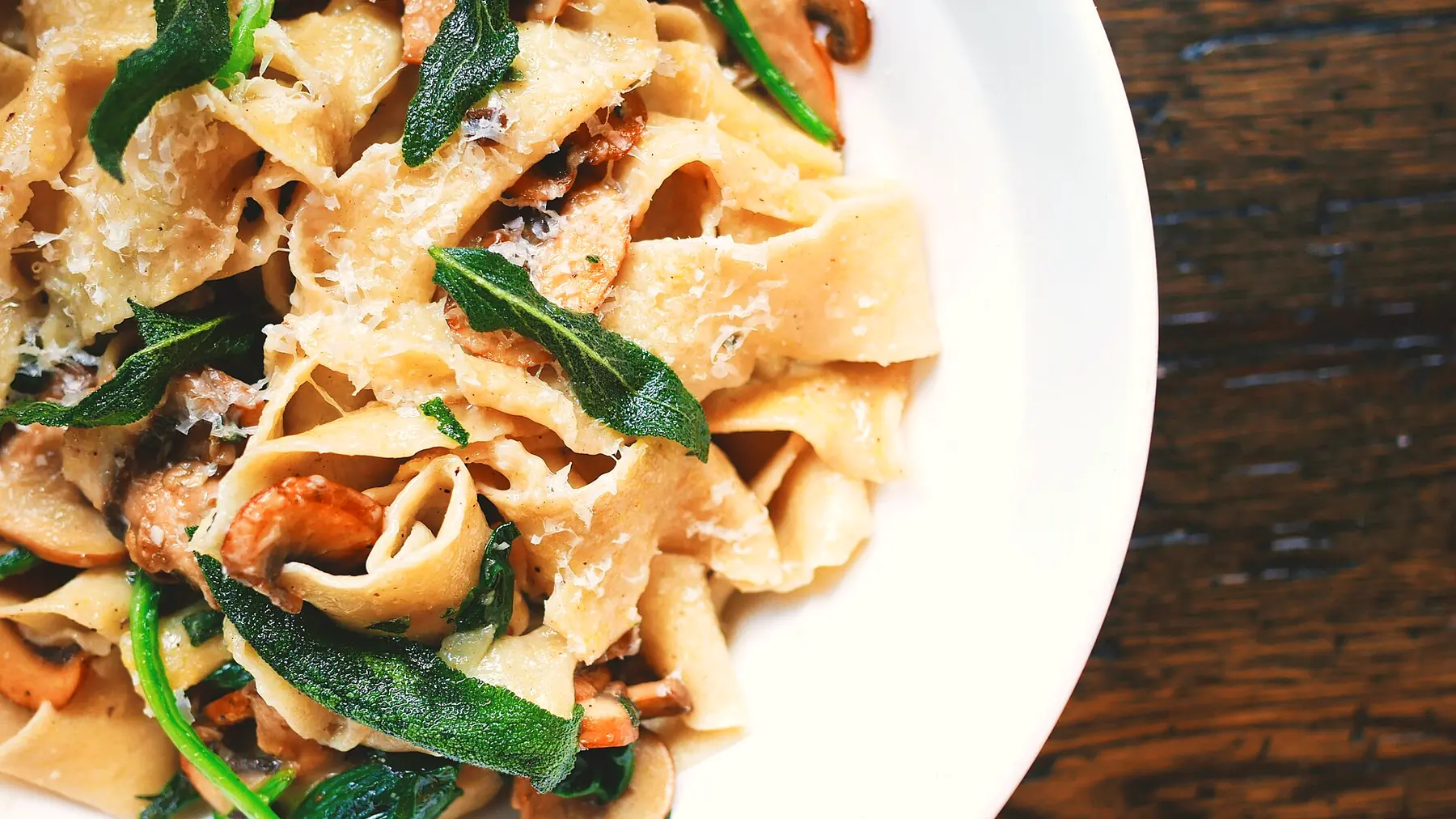 Pappardelle fatte in casa con funghi, salvia fritta e parmigiano, in un piatto bianco su un tavolo di legno scuro.