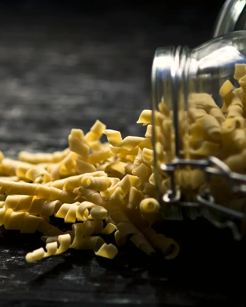 Pasta secca fatta in casa versata da un barattolo di vetro su un piano di legno scuro.